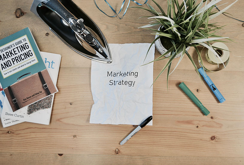 desk containing books, ironing, pens and a plant with the paper saying marketing strategy