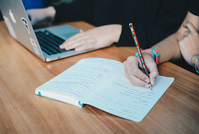 arms of a student using apple laptop while another writes in a notbook