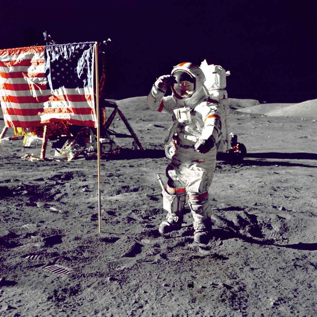 A NASA astronaut standing on the moon next to an American flag.