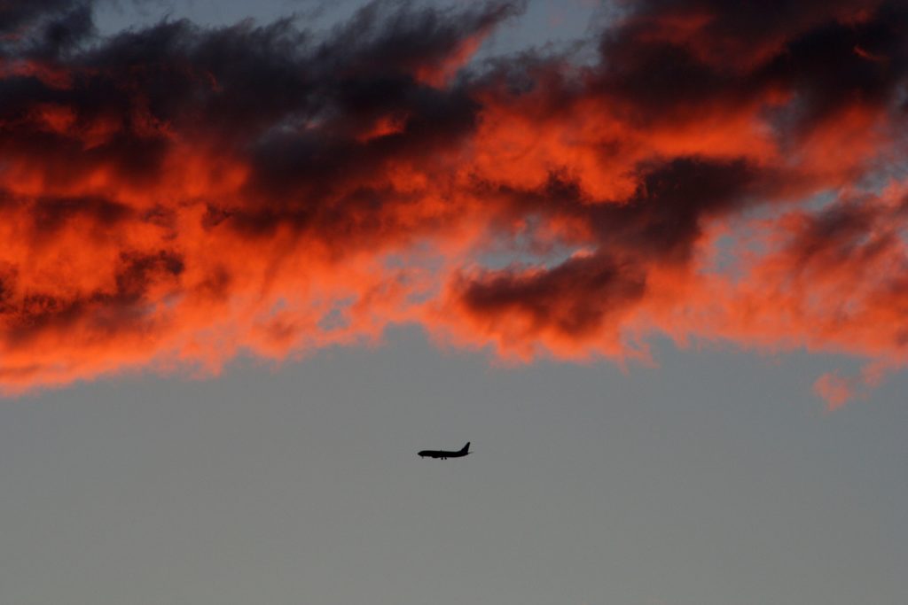 A silhouette of a plane in flight set against an orange sky with clouds.