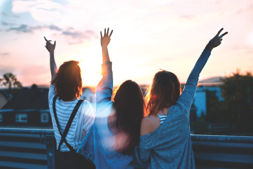 Three young women facing the sunset with their hands in the air.