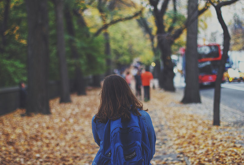 A young student walking down the road with a blue school bag on her back.