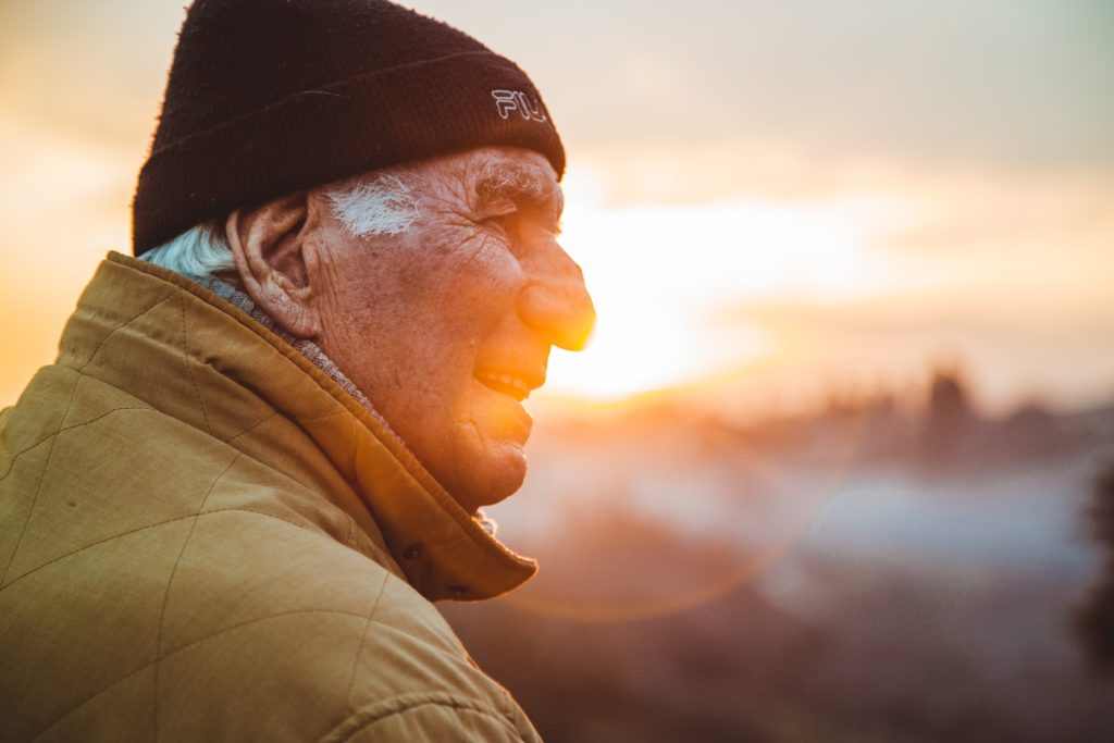 A close up portrait of an elderly man with a black beanie on with the sunset in the background.