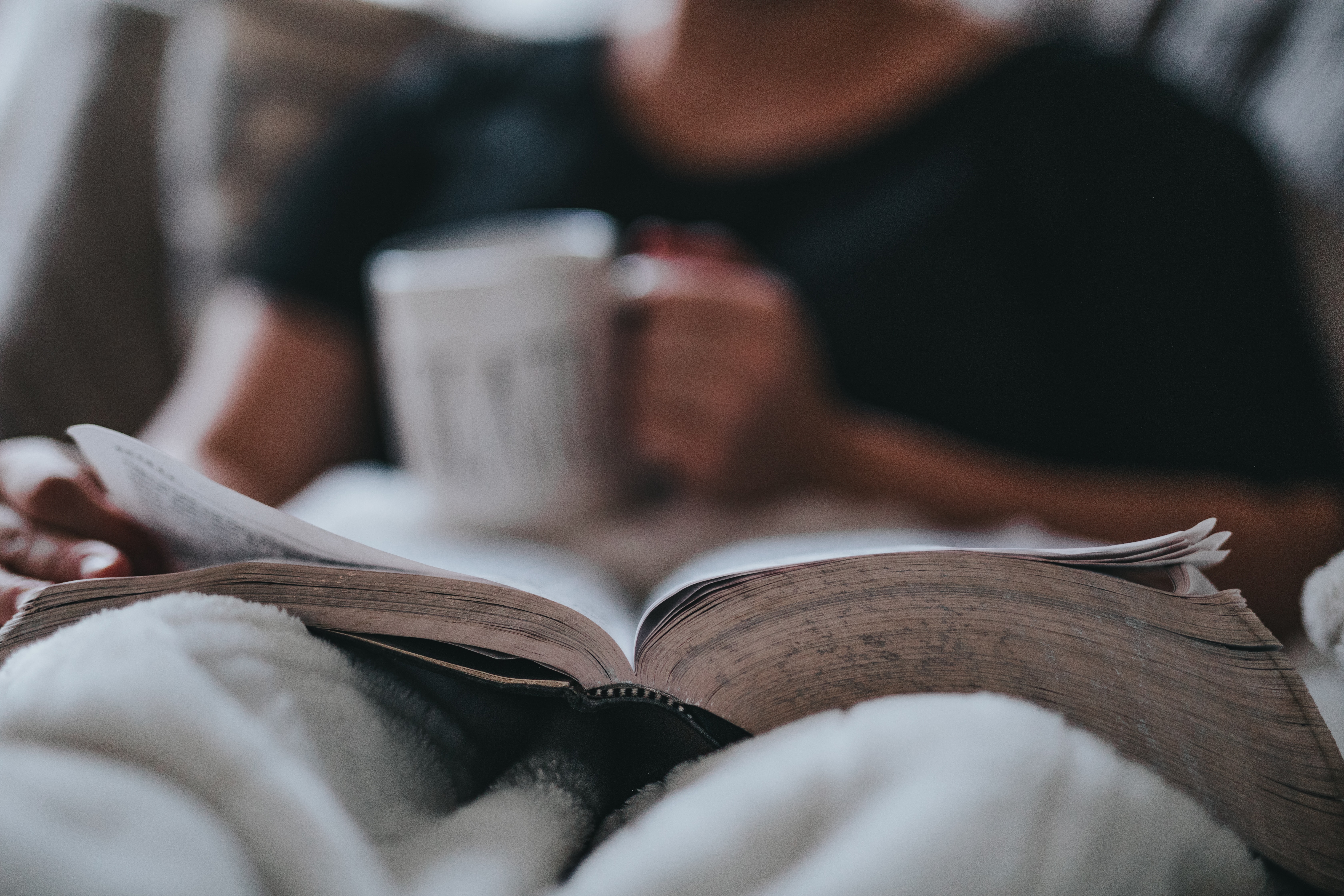 A person reading a book in bed with a cup of coffee.
