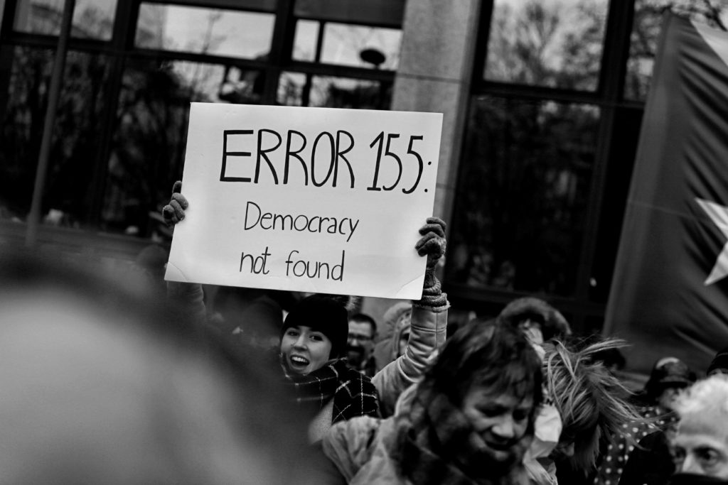A group of protestors, with one protestor holding a placard that says "Error 155: Democracy not found."