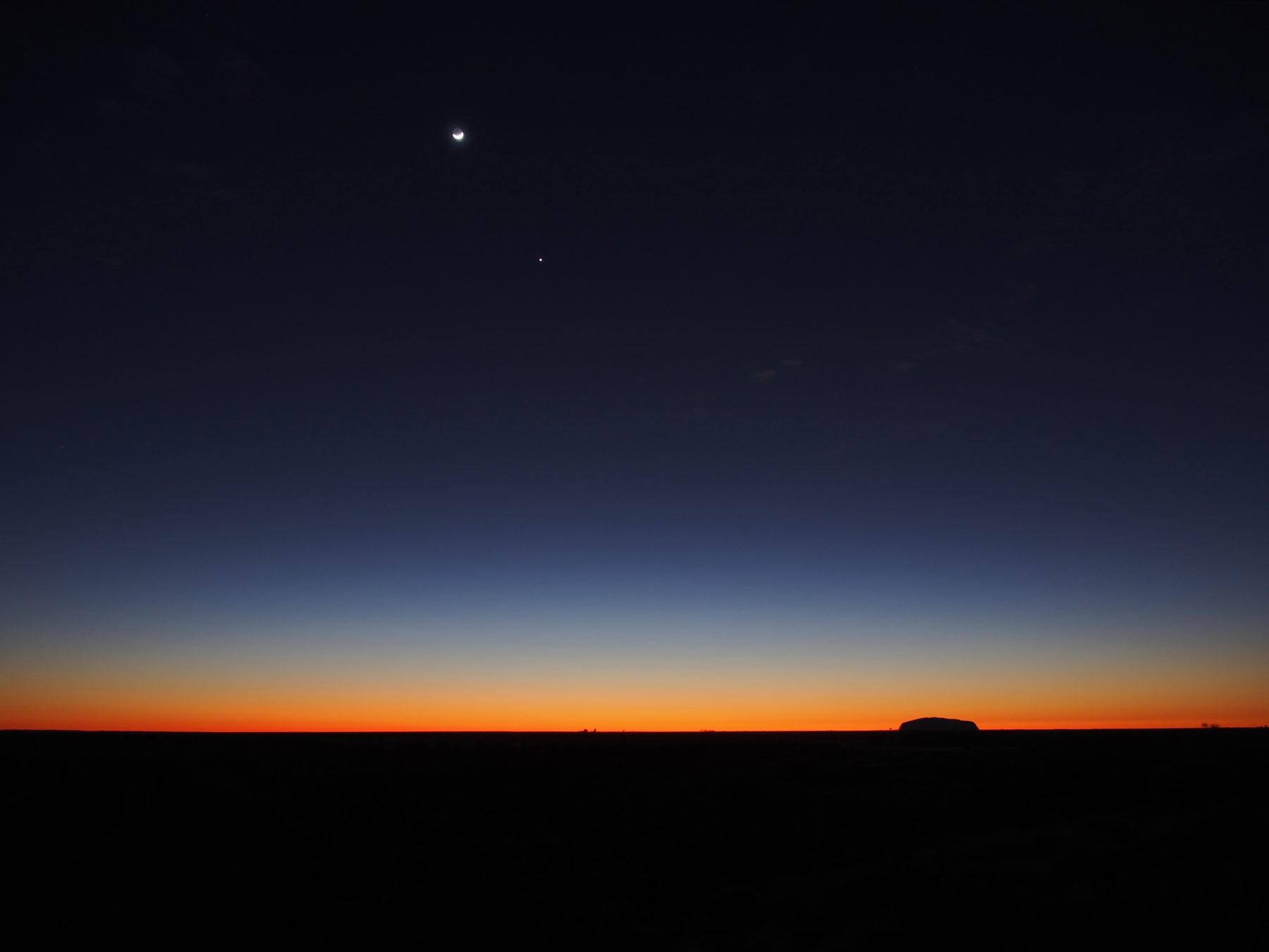 A twilight image of Uluru and the surrounding landscape with the moon and stars in the sky.