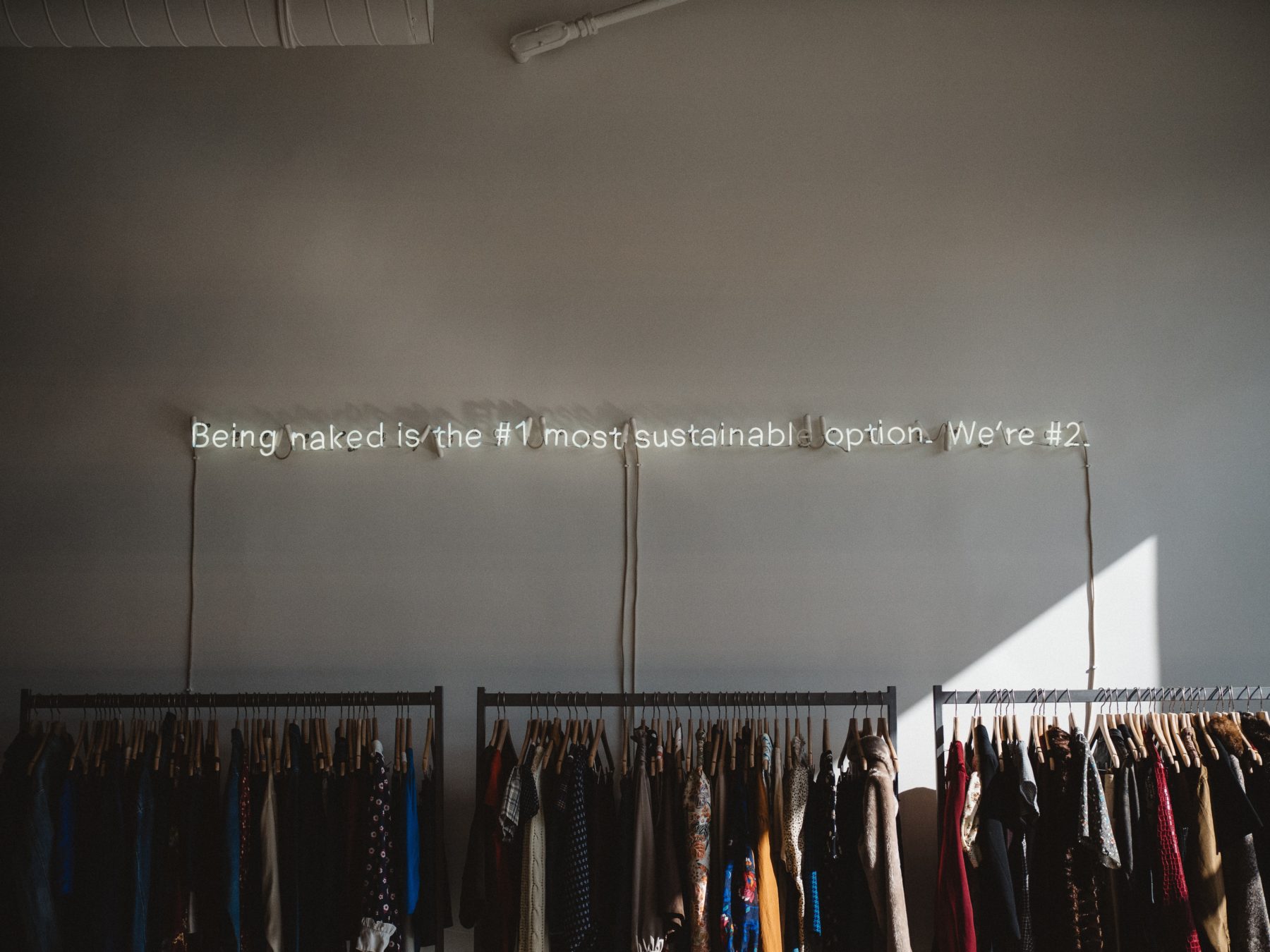 A rack of clothes with a neon sign on the wall above that says "being naked is the number one most sustainable option. We're number 2."