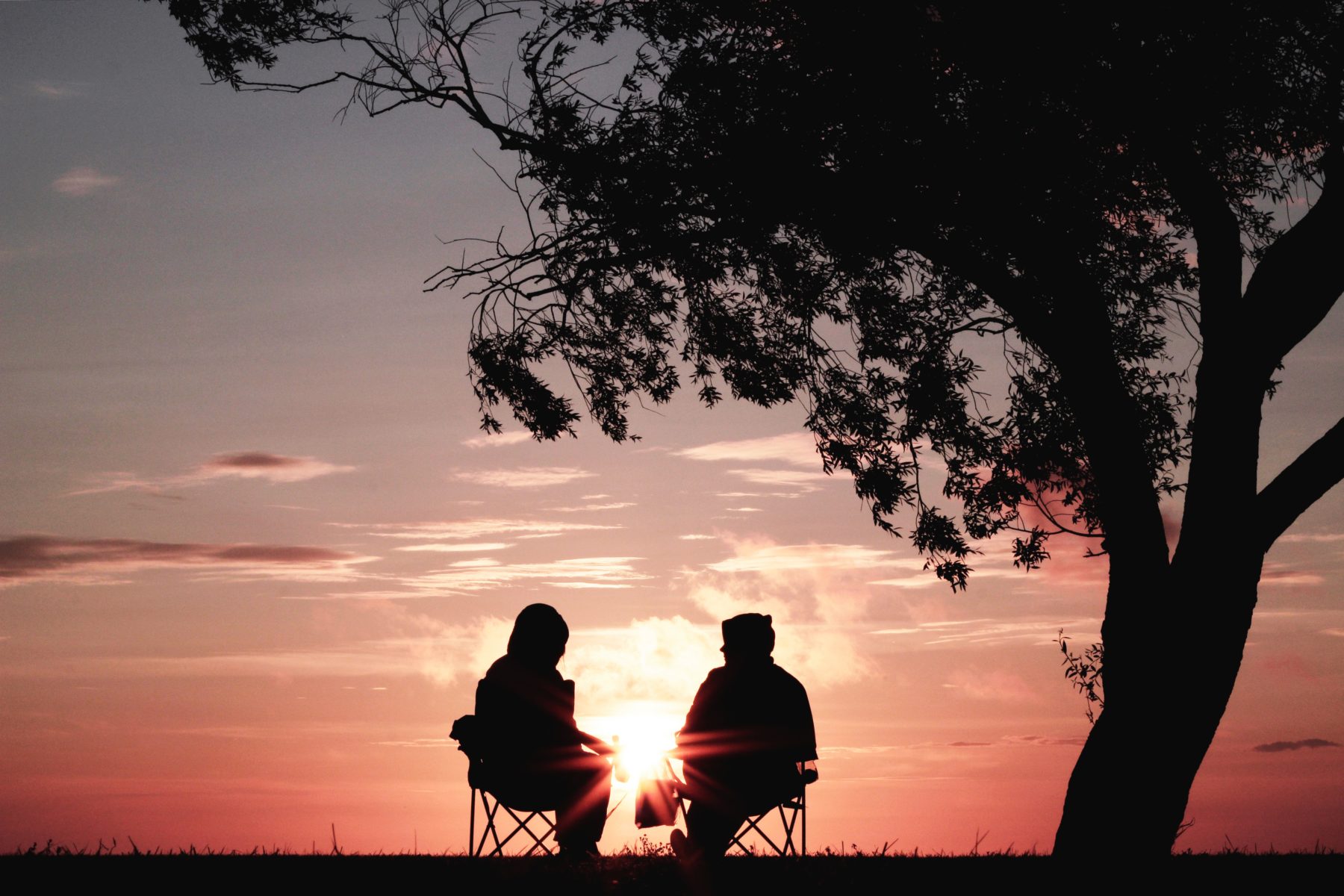 An elderly couple sitting under a tree on chairs as the sun sets in the background.