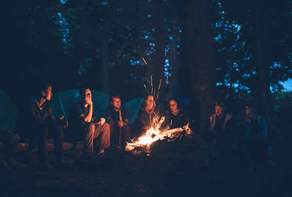 Friends sitting by the fire on a wooden log in the woods.