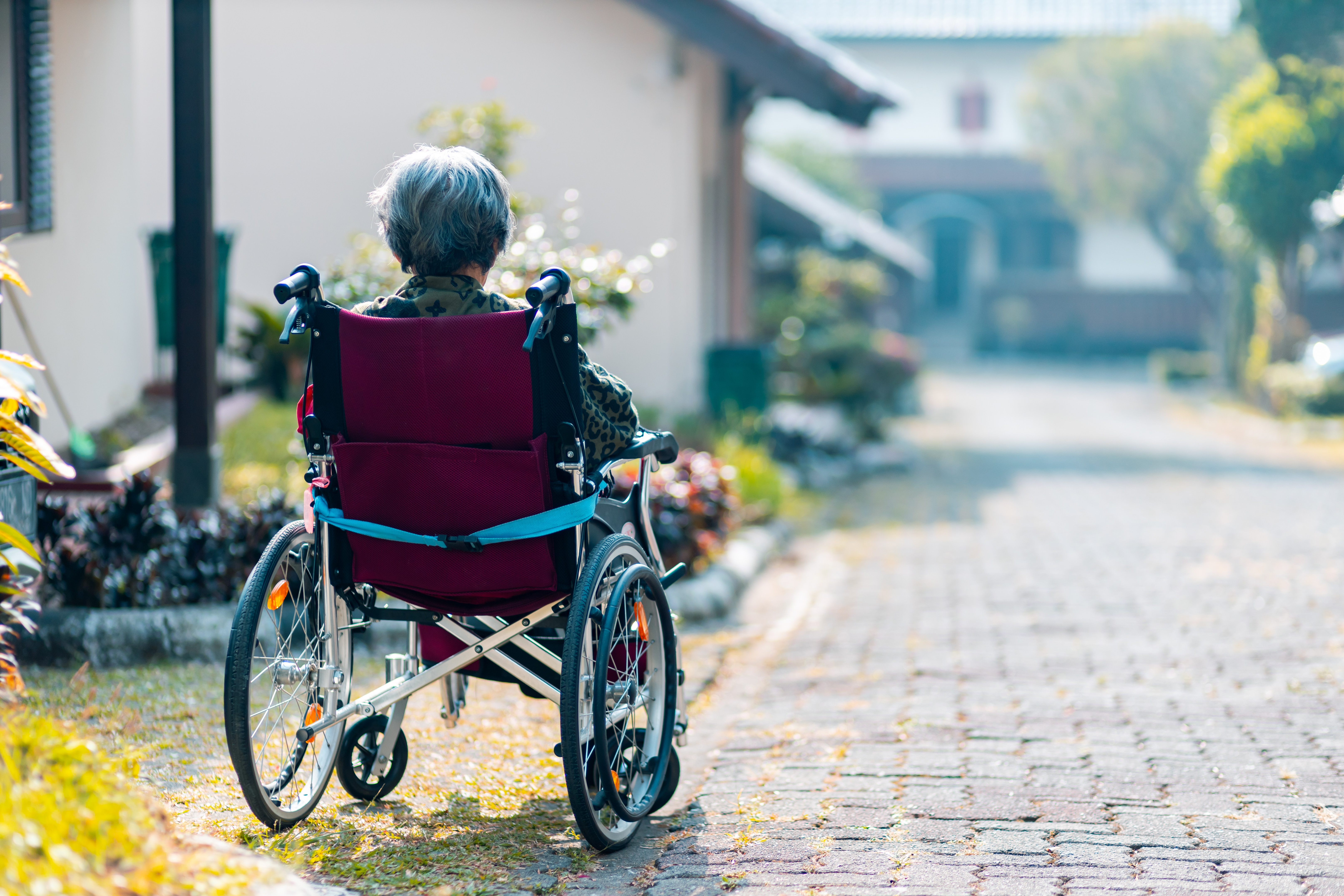 An elderly lady sitting in a wheelchair in the driveway of her home.