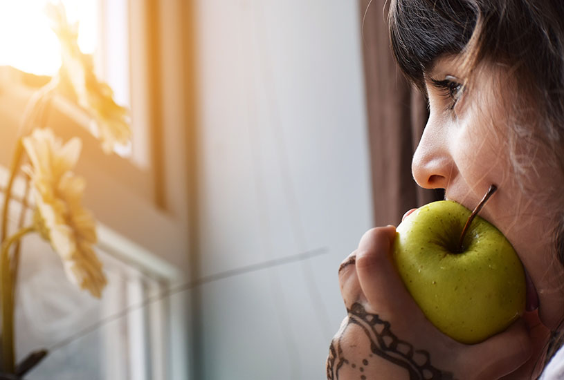 A young wmoan biting into a green apple as she gazes out of a window.