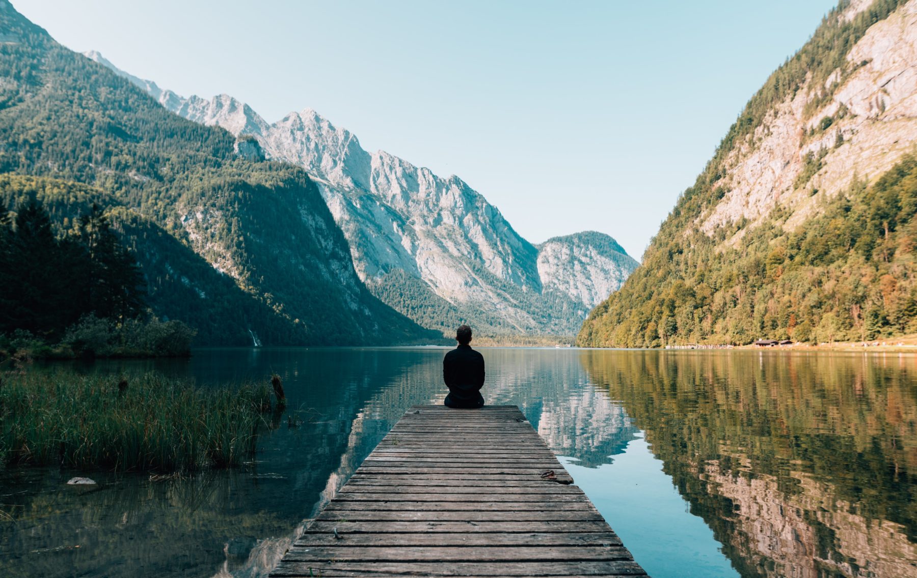 Image of man sitting on the edge of a boardwalk, surrounded by mountains and clear water  