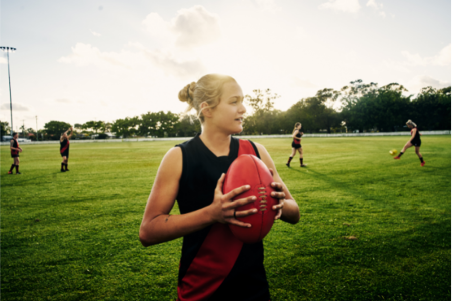 Image of a female football player. She is staying on a vivid green oval, looking to the right of the camera. She has blonde hair pulled back into a bun, and is holding a football. She is wearing a black football guernsey with a red diagonal stripe