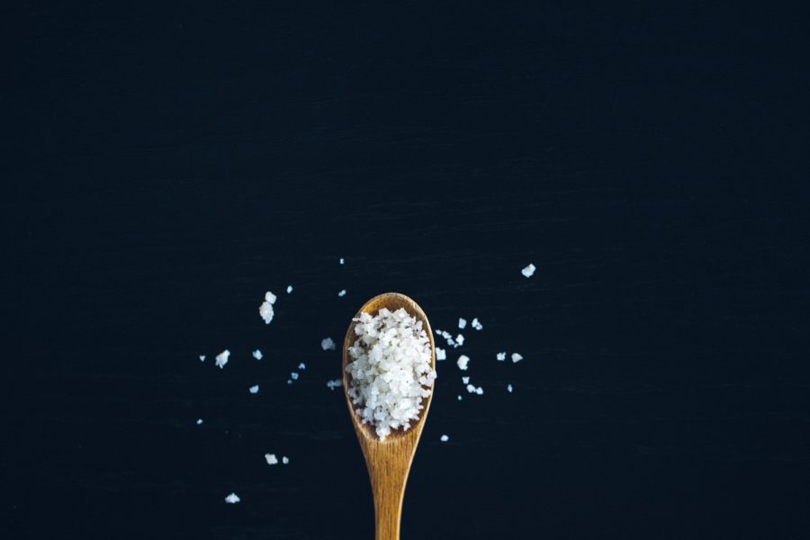 Image of a wooden spoon on a black background. The spoon is filled with flakes of white salt, and some have spilled over the sides and onto the surface it's sitting on