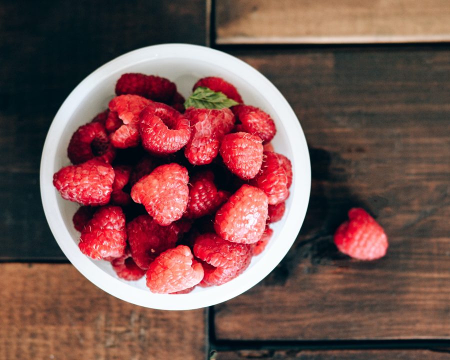 Image of a bowl of bright red raspberries, sitting on a wooden table. One of the raspberries has fallen out of the bowl and is resting on its side