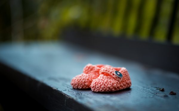A pair of pink crocheted baby shoes with embroidered blue flowers on the buckles sit on a grey park bench